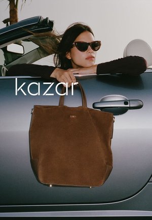 Sac fourre-tout en daim marron avec deux poignées, forme structurée et accents métalliques. Logo affiché à l'avant, placé contre une voiture.