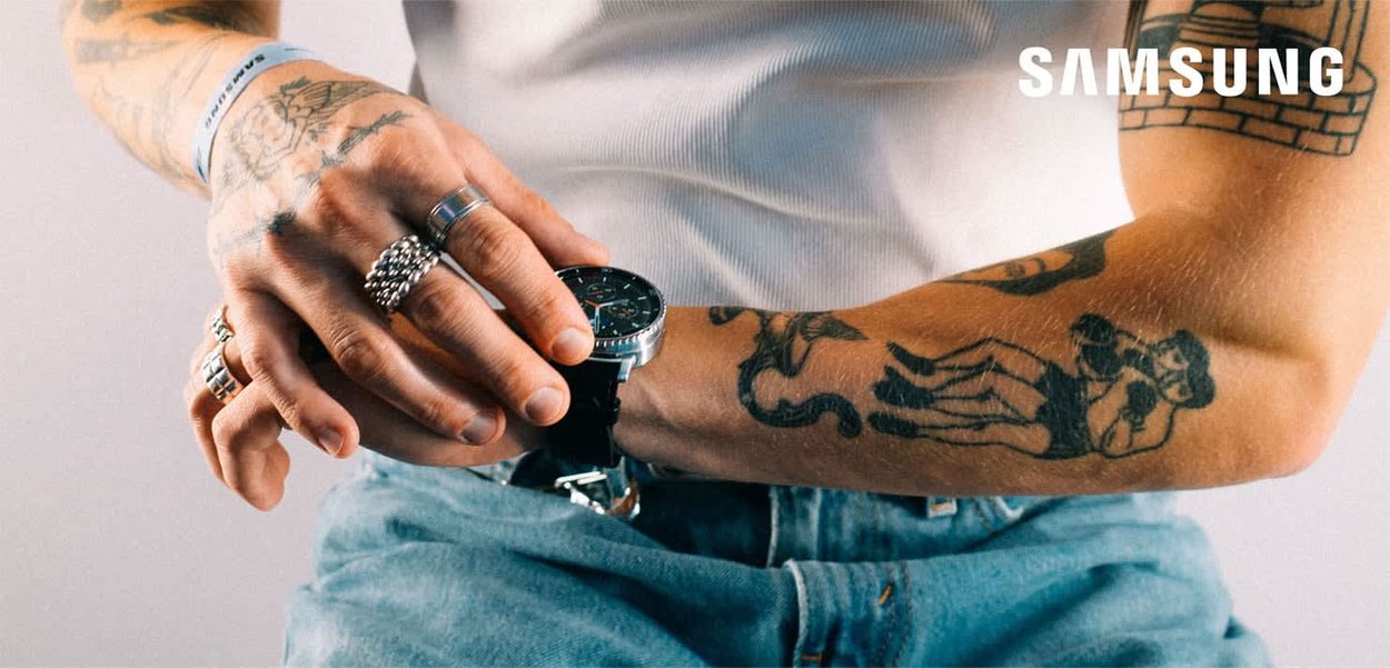 Samsung-Smartwatch mit schwarzem Armband und rundem Zifferblatt, mit silbernen Akzenten und dunklem Ziffernblatt, getragen an einem tätowierten Unterarm.