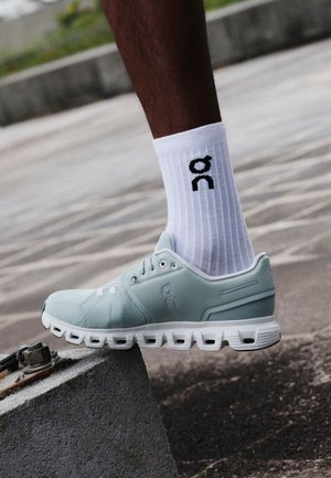 Gamba con calzini bianchi a metà polpaccio con logo e scarpa sportiva grigio chiaro che calpesta un blocco di cemento inclinato all'aperto.