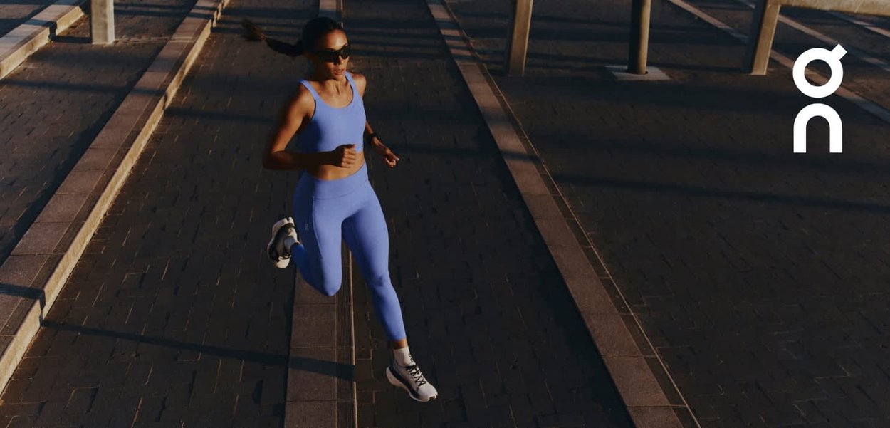 Donna in abbigliamento sportivo blu e occhiali da sole che corre su un percorso urbano in pietra illuminato dal sole, con ombre e un logo bianco a destra.