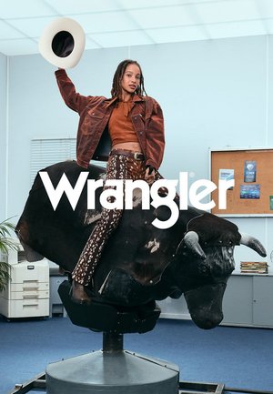 Wrangler spijkerjas, bruin, gedragen met een roestkleurig top en een gebloemde broek in bruin op een mechanische stier in een binnenomgeving.