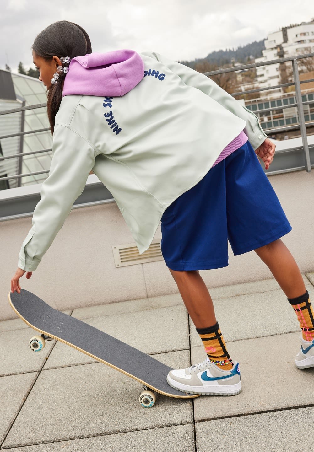 Light green oversized jacket with purple hood, blue shorts, white sneakers with teal accents, and patterned socks, skateboarding on a flat surface.