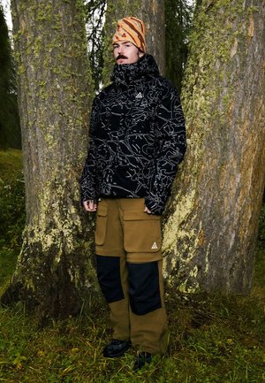 Uomo con giacca nera decorata, pantaloni cargo marroni e berretto a righe arancioni che sta in piedi tra alberi coperti di muschio in una foresta.