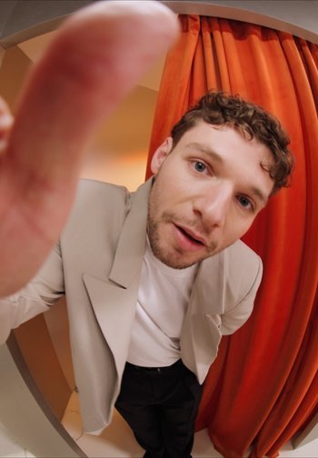 Homme en blazer beige et chemise blanche tendant la main vers un objectif fisheye, debout devant des rideaux orange dans une cabine d'essayage.