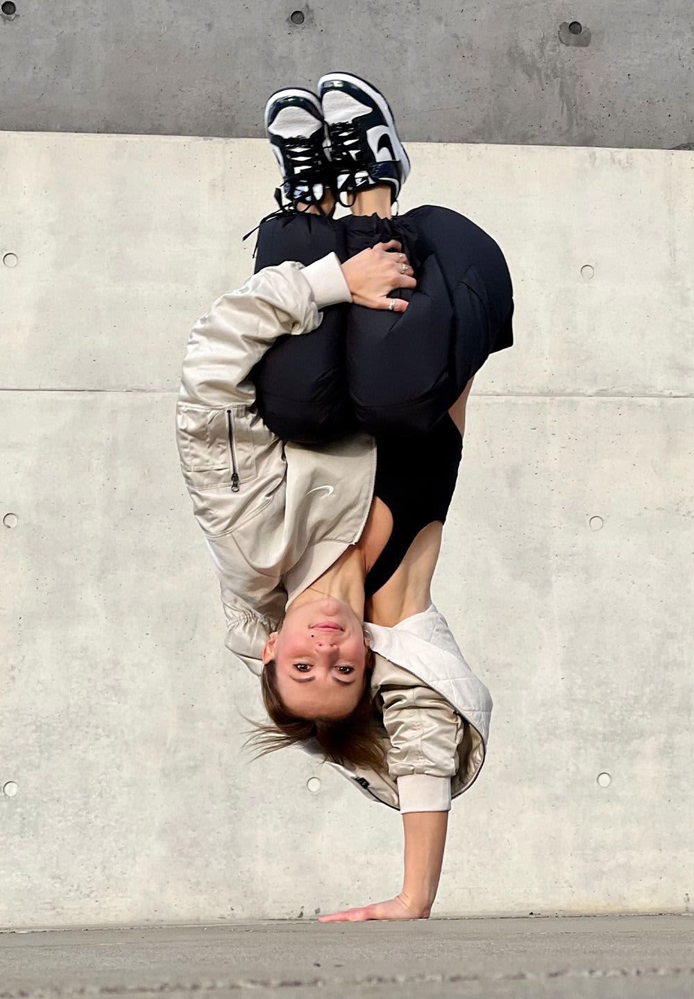 Athletic figure in a light beige bomber jacket and black leggings, performing a handstand on a concrete surface, wearing black and white sneakers.