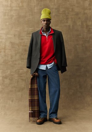 Zwarte blazer, rode pullover, blauwe shirt en losse denim jeans. Olijfgroene beanie en een bruine sjaal met ruitpatroon. Bruine veterschoenen met dikke zolen.