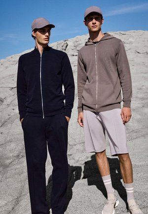 Due uomini sono in piedi su un terreno roccioso, indossano giacche con zip, cappellini, pantaloni scuri e pantaloncini chiari con scarpe da ginnastica, sotto un cielo azzurro e limpido.