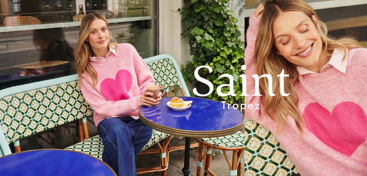 Frau in einem rosa Pullover mit Herz sitzt an einem Café-Tisch, hält eine Tasse und lächelt, auf dem Tisch liegt ein Gebäck.