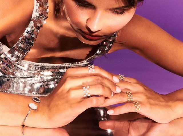 Ασημί τοπ με παγιέτες, κοντινό πλάνο χεριών στολισμένων με πολλαπλούς γεωμετρικούς δακτυλίους και ένα βραχιόλι, να αντανακλούν το φως σε μια γυαλιστερή επιφάνεια.