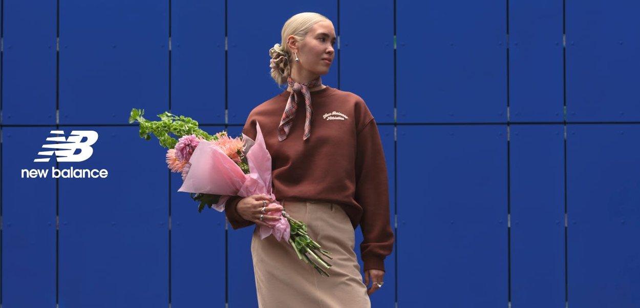 Sweat-shirt rouge brique avec logo blanc, jupe beige. Mannequin tenant un bouquet de fleurs roses devant un mur bleu.