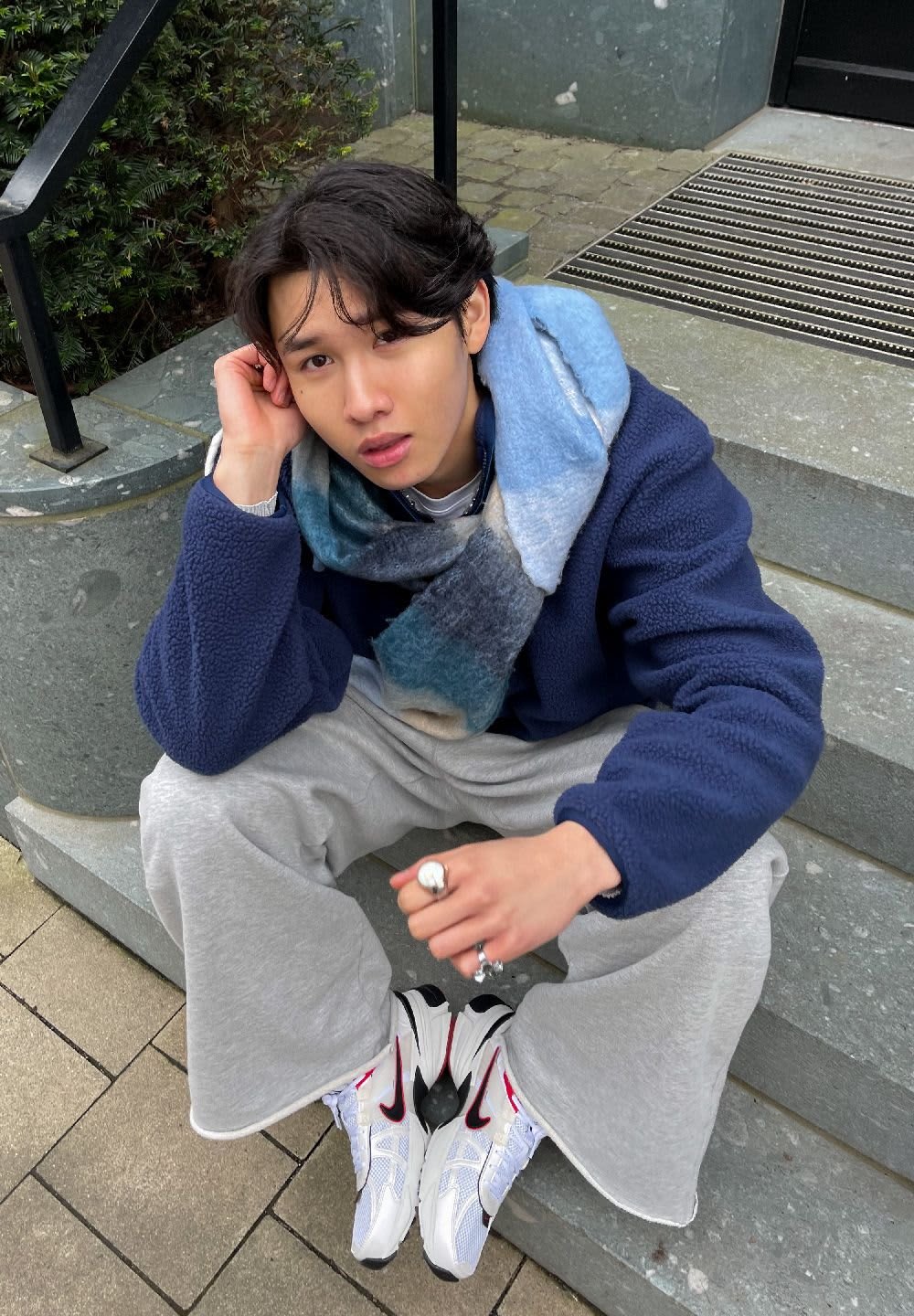 Person seated on stone steps, wearing a navy fleece jacket, grey sweatpants, and white sneakers with black accents. Multicolored scarf draped.