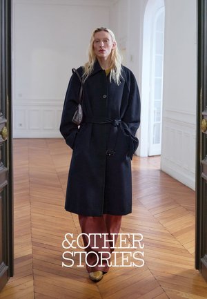 Kvinde iført lang sort frakke med bælte, brune bukser og mønstrede sko står i et lyst rum med trægulv i sildebensmønster og hvide vægge.