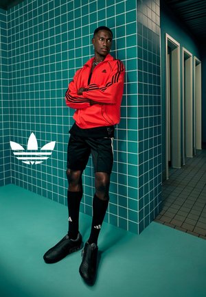Rode adidas zip-up jas met zwarte strepen, zwarte shorts en stijlvolle zwarte schoenen. Geplaatst tegen teal betegelde muren en vloer.