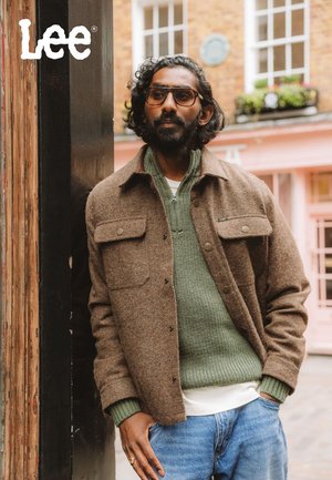 Brown wool-blend shirt jacket with chest pockets, green knitted sweater underneath, denim jeans, and light-colored t-shirt.