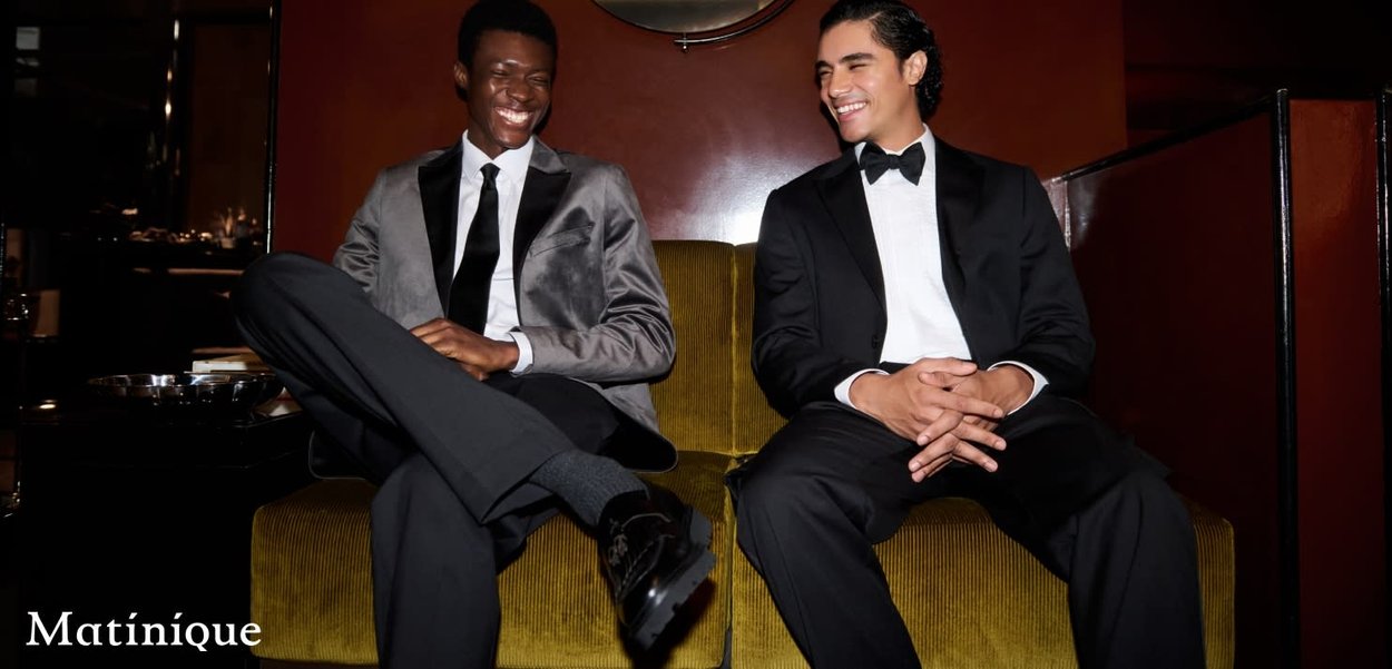 Zwei Männer sitzen auf einer grünen Samtcouch. Der eine trägt einen grauen Smoking mit Schalkragen; der andere einen schwarzen Smoking mit Fliege.