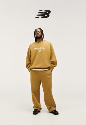 Homme aux cheveux tressés portant un sweat-shirt New Balance beige et un pantalon assorti avec des baskets noires, logo New Balance au-dessus.