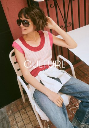 Frau mit kurzen braunen Haaren und Sonnenbrille sitzt auf einem weißen Stuhl an einem Tisch, trägt ein pink-rotes ärmelloses Oberteil und blaue Jeans.