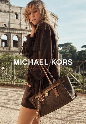 Brown leather handbag with a patterned surface, short handles, and metal accents. Model wears a fringed brown suede outfit, posed outdoors.