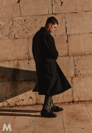 Manteau en laine noir au fit ample, superposé sur un pantalon gris et des chaussures noires brillantes. Adossé à un mur en pierre texturé.