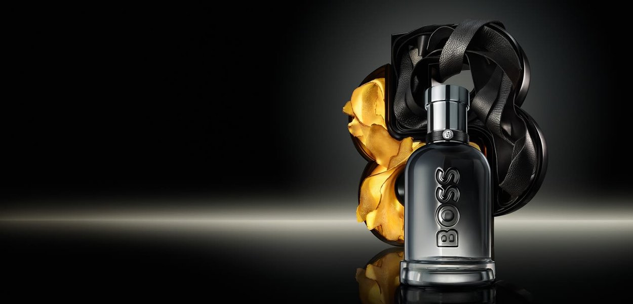 Bouteille de parfum en verre noir avec "BOSS" gravé. Entourée d'un ruban noir texturé et d'un accent floral jaune, sur un fond sombre.