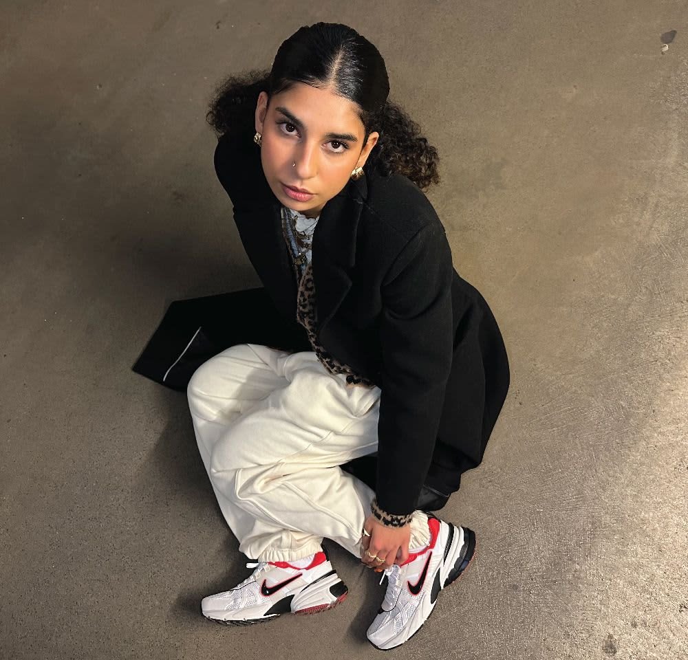 White oversized sneakers with red accents and black swoosh, worn with loose cream trousers and a black coat, featuring a stylish pattern beneath.
