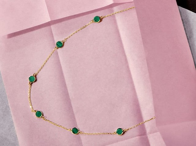 Λεπτή χρυσή αλυσίδα με έξι ομοιόμορφα κατανεμημένες στρογγυλές πράσινες πολύτιμες πέτρες σε ροζ και γκρι φόντο από διπλωμένο χαρτί.