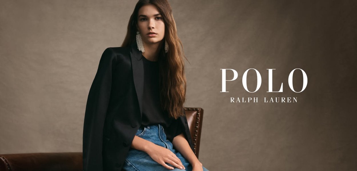 Blazer noir sur un haut noir, associé à une jupe en denim bleu. Cheveux longs et ondulés. Le texte « POLO RALPH LAUREN » en blanc. Fond marron.