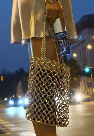 Person wearing a beige dress holds a sparkling gold handbag and a blue bottle labeled "Sol Janeiro 76" on a city street at night.