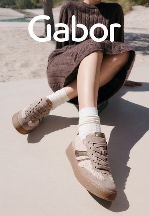 Beige lace-up sneakers with a mix of suede and textured fabric, featuring a rubber sole and subtle metallic accents. White ribbed socks visible.