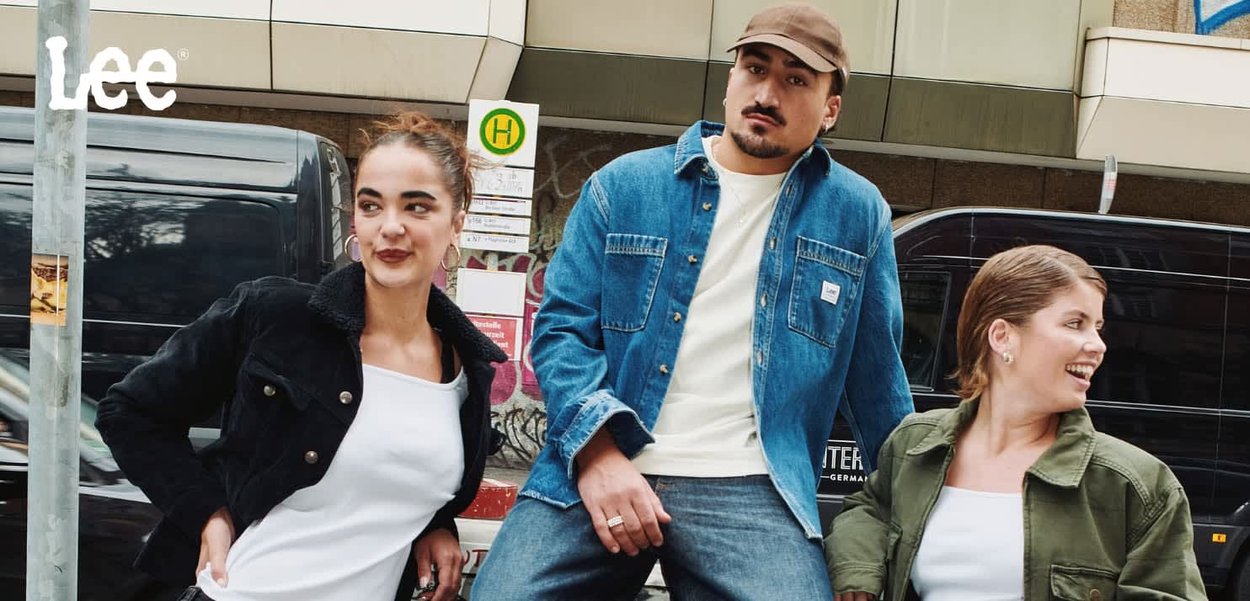 Three individuals pose outdoors. Two wear white tops, one in a black cord jacket, another in an olive green jacket. The third wears a denim jacket.