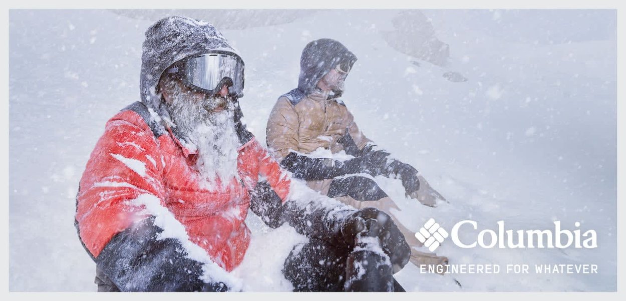 Zwei Personen sitzen in einer verschneiten Umgebung, eine in einer leuchtend roten, isolierten Jacke, die andere in einer tanfarbenen Kapuzenjacke, beide in Winterbekleidung.