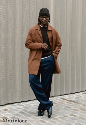 Cappotto impermeabile marrone sopra un maglione scuro, abbinato a pantaloni blu navy. Completa il look un berretto nero e occhiali. Sfondo grigio testurizzato.