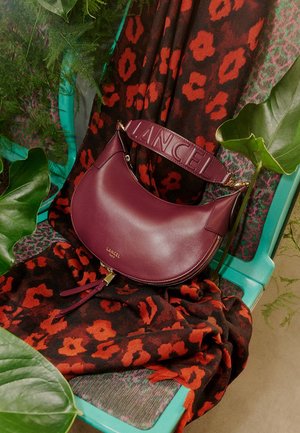 Borsa in pelle bordeaux con forma curva, tracolla con la scritta "LANCEL" in rilievo, dettagli con cerniera e accentuazioni a nappina su uno sfondo di tessuto a motivo.