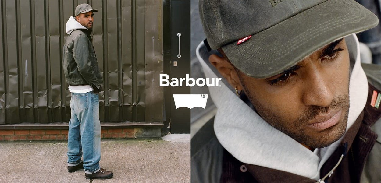 Giacca Barbour verde scuro, sovrapposta a un felpa grigia con cappuccio. Jeans blu e stivali scuri. Cappellino grigio con dettaglio del logo. Sfondo urbano.