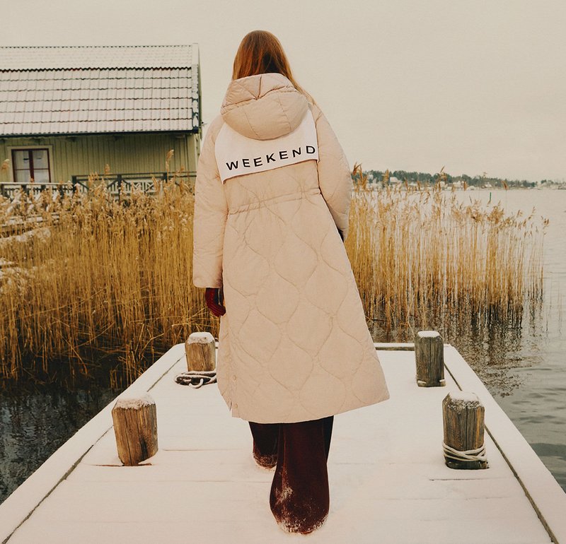 Beige vadderad vinterkappa med huva, med ordet "WEEKEND" i svart över ryggen. Bärs över mörka, löst sittande byxor.