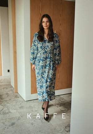Blaues, blumiges Midikleid mit langen Puffärmeln, verziert mit weißen Mustern. Getragen mit schwarzen spitzen Schuhen, vor einem neutralen Hintergrund.