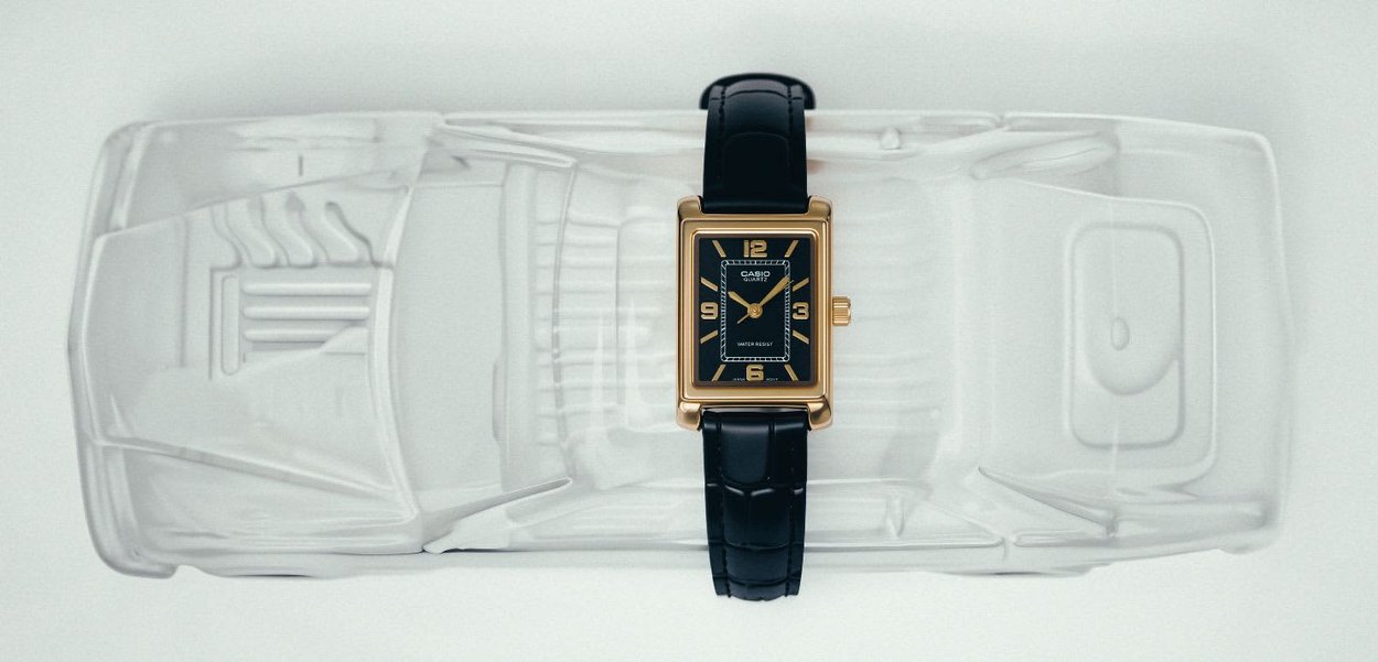 Reloj de pulsera Casio rectangular dorado con correa de cuero negro colocado sobre un modelo de coche de plástico translúcido visto desde arriba.
