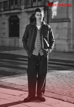 Model trägt eine dunkelgraue Jacke über einem karierten Hemd, kombiniert mit schwarzen Hosen und Schuhen. Die Kulisse wirkt urban mit Kopfsteinpflaster.