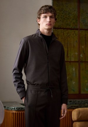 Graue Zip-Jacke mit hohem Kragen, kombiniert mit passenden Hosen mit elastischem Bund. Glatte Stoffstruktur mit einem entspannten Schnitt.