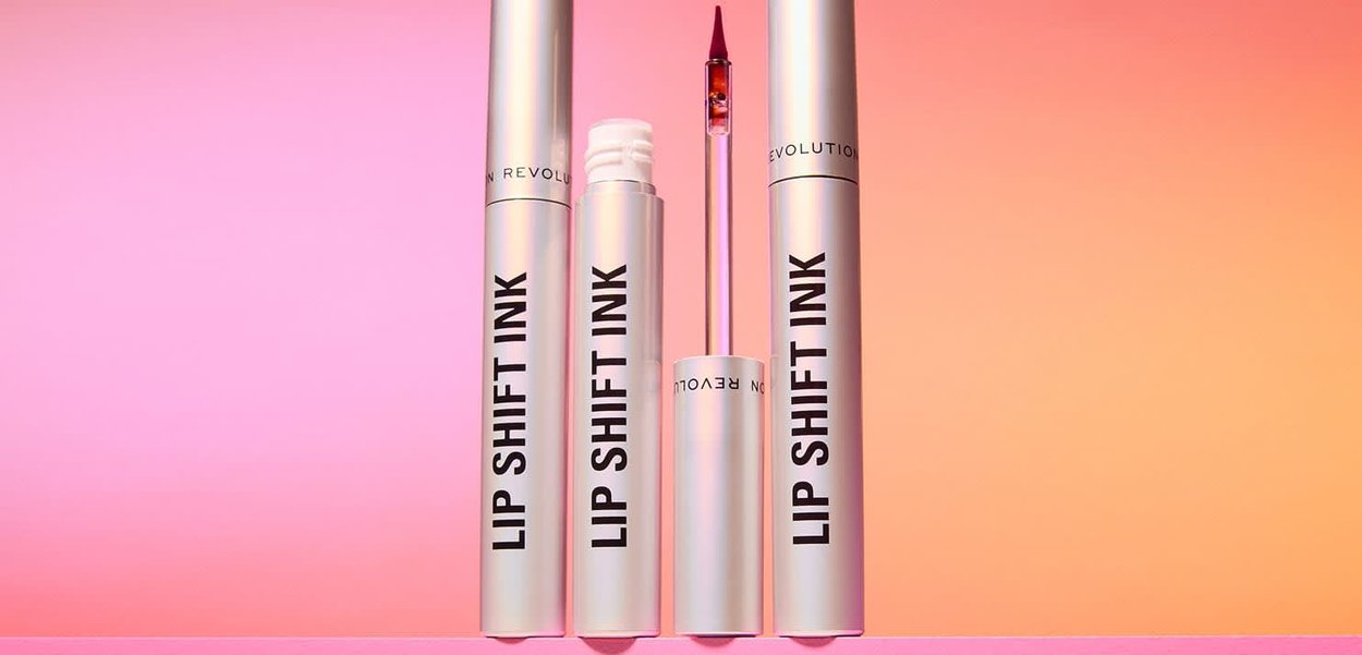 Tre tubi bianchi di rossetto "Lip Shift Ink" sono disposti in verticale su una superficie rosa con uno sfondo sfumato rosa-arancione, uno con l'applicatore esposto.