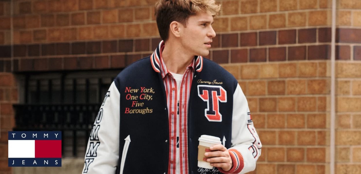 Chaqueta varsity de Tommy Jeans en azul marino y blanco, con acentos rojos y texto bordado. Modelo sosteniendo una taza de café, de pie frente a una pared de ladrillo.