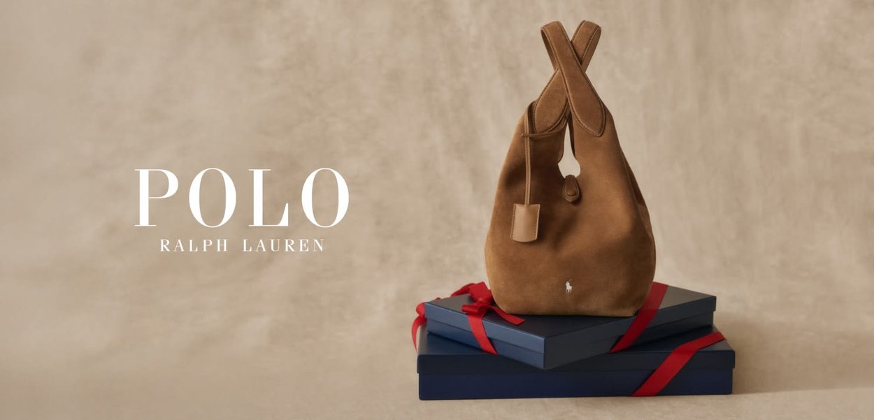 Bolso hobo de ante marrón con dos asas y una pequeña etiqueta, apoyado sobre dos cajas apiladas con cinta roja, sobre un fondo neutro.