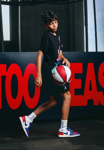 Junge Person in einem schwarzen T-Shirt mit einem bunten Grafikdesign, schwarzen Shorts und blau/roten Basketballschuhen, die einen rot/weißen Basketball dribbelt.