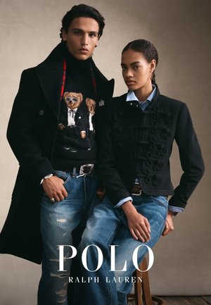 Giovane uomo con cappotto nero e maglione teddy accanto a giovane donna con giacca nera e camicia blu, entrambi con jeans blu, uno seduto e l'altro in piedi.