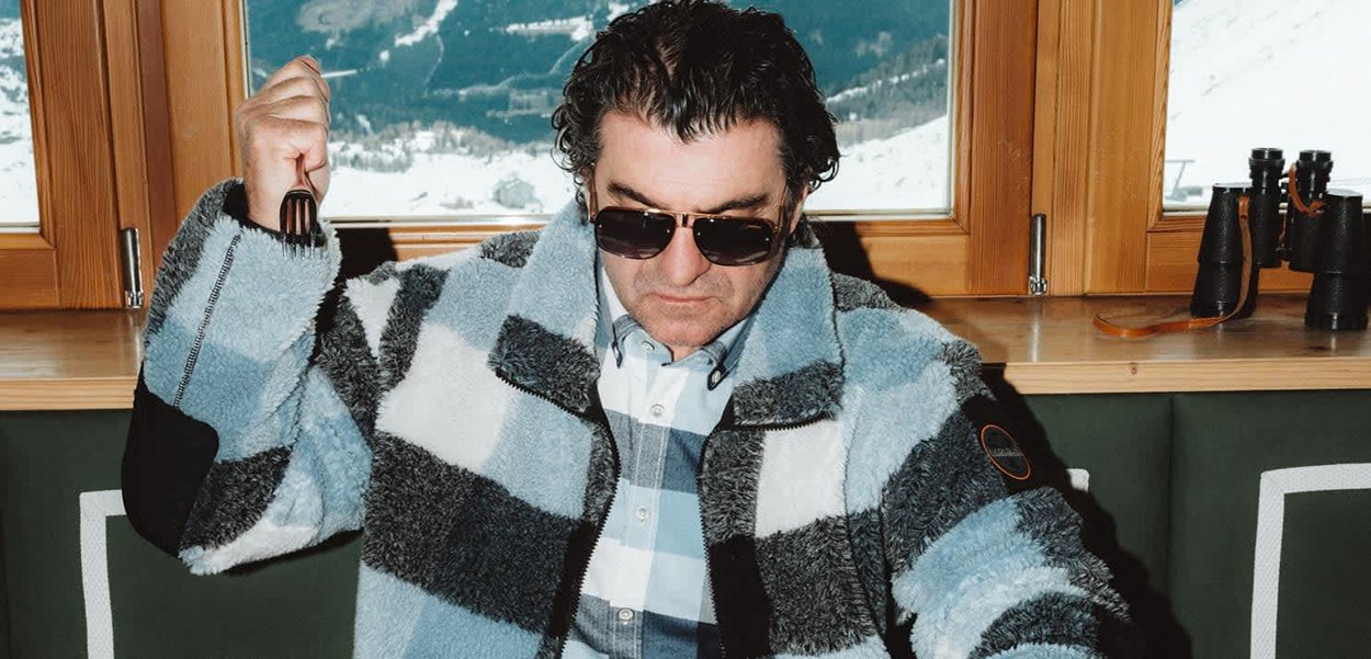 Giacca in pile sfocata a righe blu e grigie con colletto nero; chi la indossa tiene una forchetta e porta occhiali da sole davanti a uno sfondo innevato.