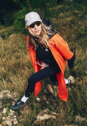 Mujer con chaqueta naranja, leggings negros, gorra blanca y gafas de sol caminando por un terreno rocoso con hierba, con árboles al fondo.