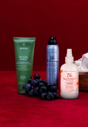 Quattro prodotti per capelli su uno sfondo rosso: crema ricci verde Aveda, spray texture blu Bumble and Bumble, e due primer Bumble and Bumble in bianco e arancione. Uva nera a sinistra.