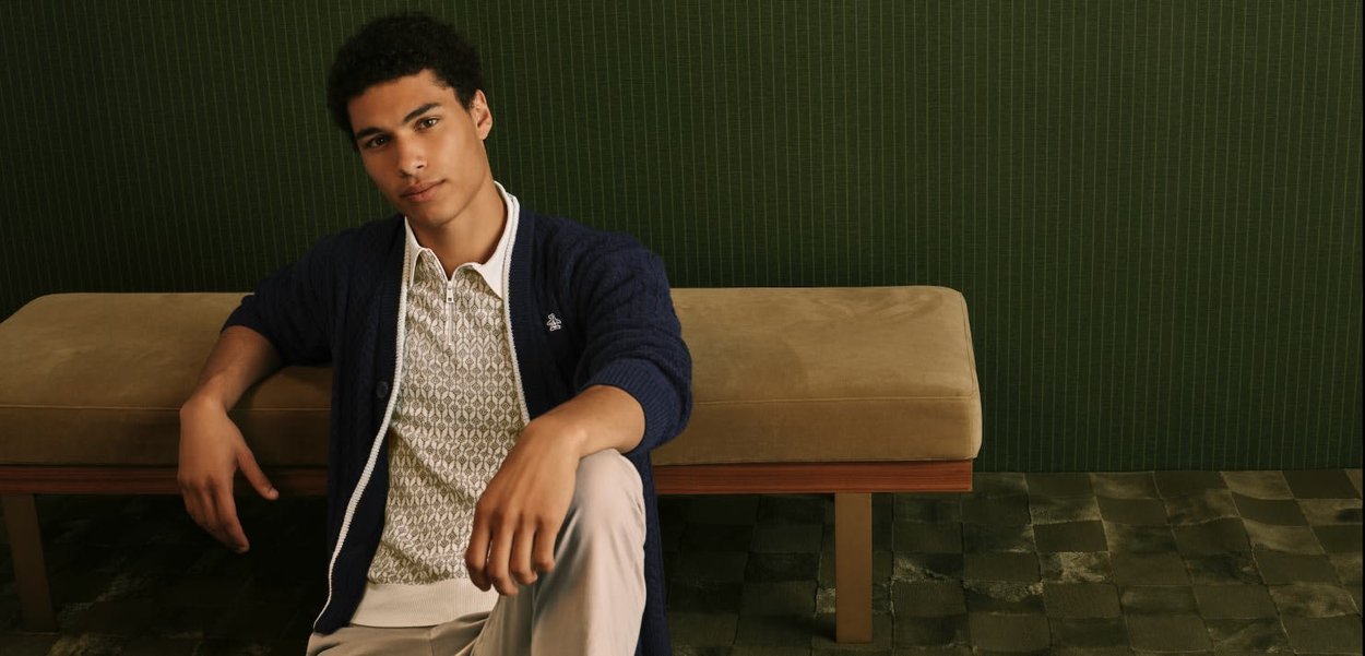 Junger Mann in hellgemustertem Hemd, marineblauer Strickjacke und beigen Hosen sitzt auf dem Boden und lehnt sich an eine beige Bank vor einem dunkelgrünen Wandhintergrund.