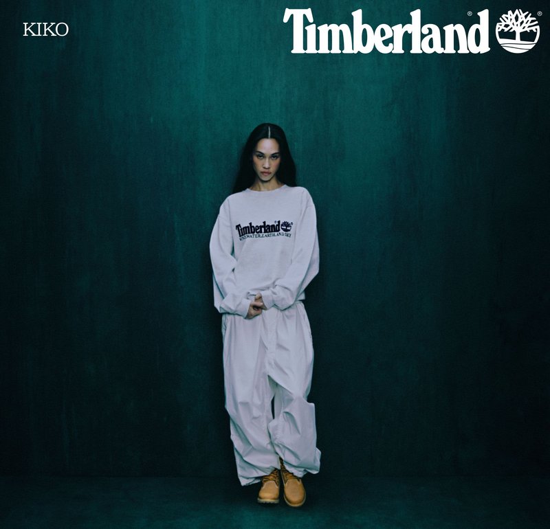 Bluza z logo "Timberland", białe luźne spodnie oraz żółte buty Timberland. Model stoi na tle ciemnozielonym.