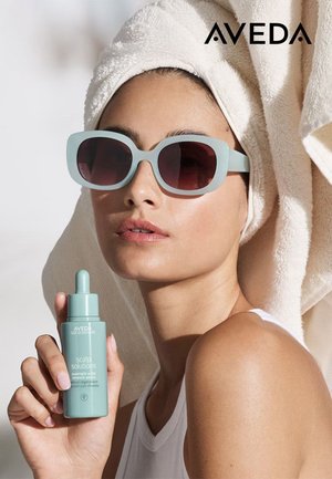 Aveda Scalp Solutions-flaske, mintgrønn, med dråpetopp, holdt av en person med store lyseblå solbriller med mørke glass, håndkle i håret.